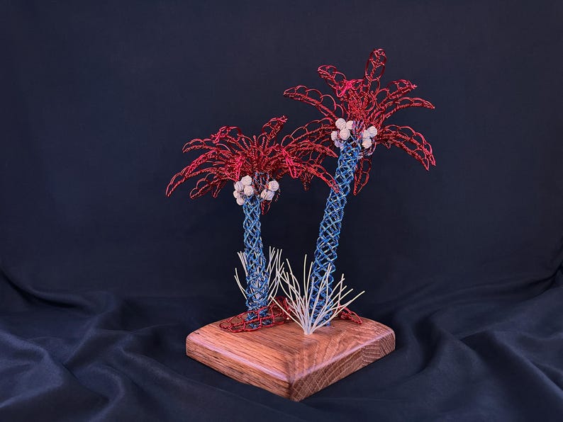 May include: A decorative sculpture featuring two palm trees crafted from twisted wire. The trunks are blue, the fronds are red, and the coconuts are white. The trees are mounted on a square wooden base with a small cluster of white wire grass.