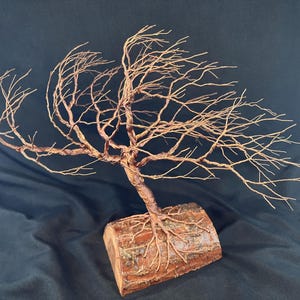 Puede incluir: Una escultura de &aacute;rbol hecha de alambre de cobre trenzado, con ramas intrincadas que se extienden hacia afuera. La base del &aacute;rbol est&aacute; anclada en una pieza cil&iacute;ndrica de madera natural con corteza visible, mostrando las ra&iacute;ces de alambre integradas.