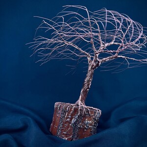 Puede incluir: Una escultura de alambre de un &aacute;rbol con un acabado de cobre met&aacute;lico, que presenta un tronco retorcido y ramas finas y extendidas. El &aacute;rbol est&aacute; anclado en una base rectangular de color marr&oacute;n texturizado que recuerda a la madera.