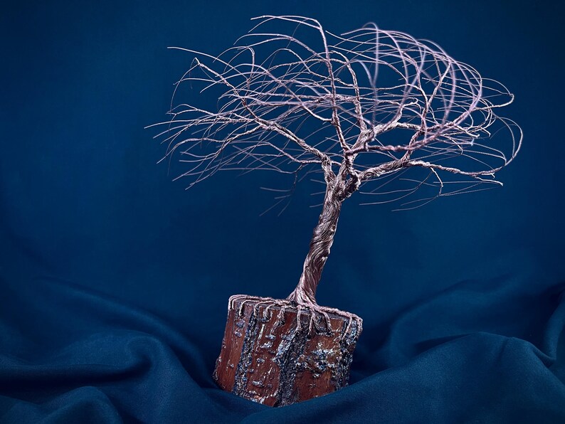 Puede incluir: Una escultura de alambre de un &aacute;rbol con un acabado de cobre met&aacute;lico, que presenta un tronco retorcido y ramas finas y extendidas. El &aacute;rbol est&aacute; anclado en una base rectangular de color marr&oacute;n texturizado que recuerda a la madera.