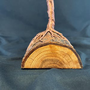 Puede incluir: Una base de madera semicircular con un exterior de corteza rugosa y texturizada y una secci&oacute;n transversal lisa de color claro que muestra anillos de crecimiento visibles. Una fina estructura de alambre de cobre trenzado se eleva.