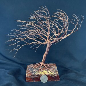 Puede incluir: Una escultura de &aacute;rbol hecha de alambre de cobre trenzado, con ramas intrincadas que se extienden hacia un lado. El &aacute;rbol est&aacute; anclado a una base de madera rectangular con corteza visible, y una moneda est&aacute; colocada en la parte frontal.