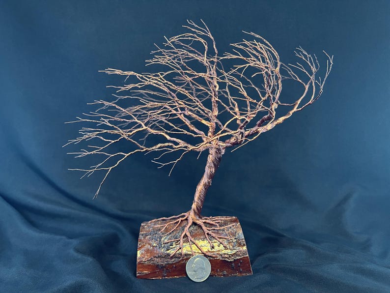 Puede incluir: Una escultura de &aacute;rbol hecha de alambre de cobre trenzado, con ramas intrincadas que se extienden hacia un lado. El &aacute;rbol est&aacute; anclado a una base de madera rectangular con corteza visible, y una moneda est&aacute; colocada en la parte frontal.