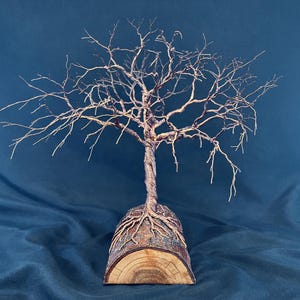 Puede incluir: Una escultura de alambre de un &aacute;rbol sin hojas con un tronco retorcido y ramas extendidas, anclada a una base de tronco de madera semicil&iacute;ndrica. El alambre met&aacute;lico es de color marr&oacute;n oscuro y la base de madera muestra vetas naturales.