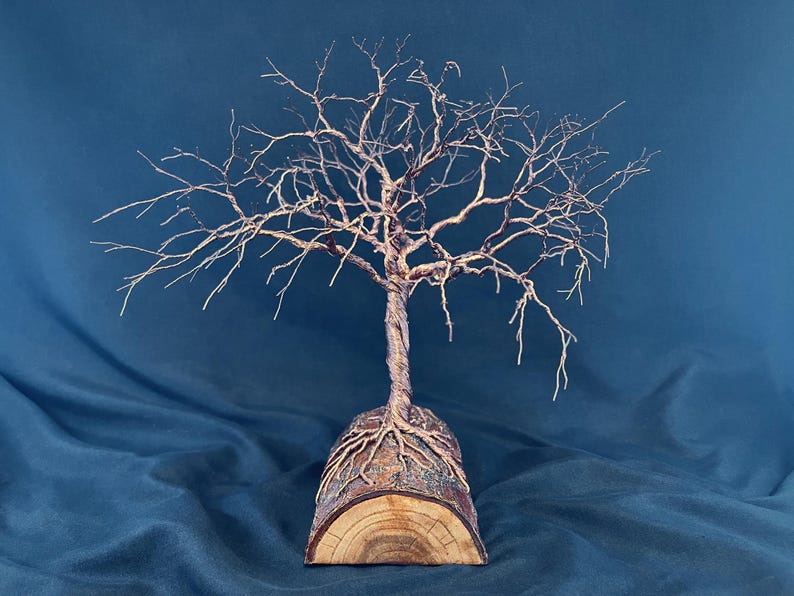 Puede incluir: Una escultura de alambre de un &aacute;rbol sin hojas con un tronco retorcido y ramas extendidas, anclada a una base de tronco de madera semicil&iacute;ndrica. El alambre met&aacute;lico es de color marr&oacute;n oscuro y la base de madera muestra vetas naturales.