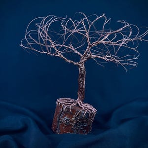Puede incluir: Una escultura de alambre de un &aacute;rbol con acabado en cobre met&aacute;lico, que presenta un tronco retorcido y ramas finas y extendidas. La base del &aacute;rbol est&aacute; anclada en un bloque de madera cil&iacute;ndrico con textura de corteza visible.