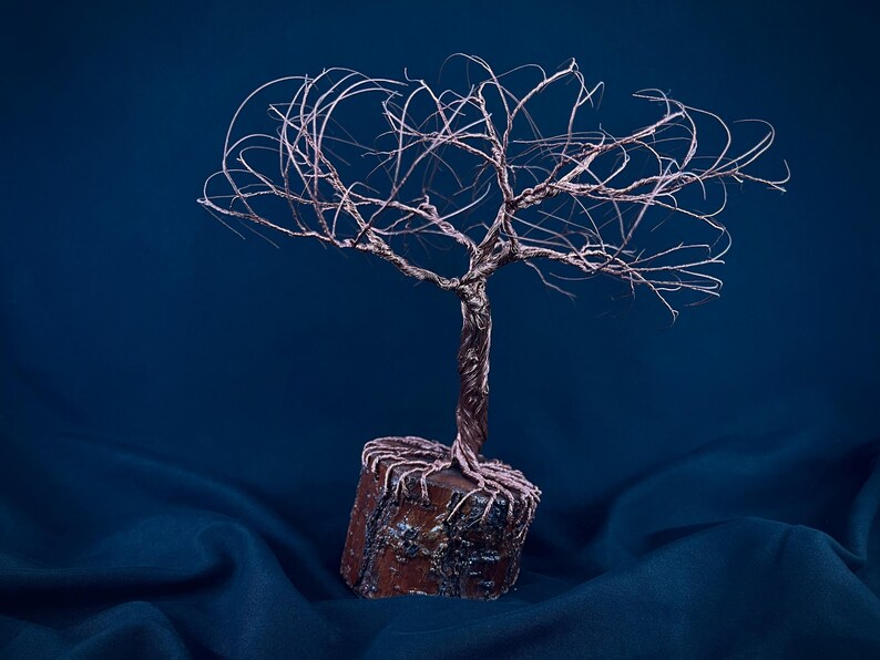Puede incluir: Una escultura de alambre de un &aacute;rbol con acabado en cobre met&aacute;lico, que presenta un tronco retorcido y ramas finas y extendidas. La base del &aacute;rbol est&aacute; anclada en un bloque de madera cil&iacute;ndrico con textura de corteza visible.