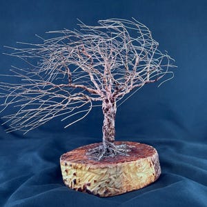 May include: A wire sculpture of a tree with thin, metallic branches extending to one side as if blown by the wind. The trunk is formed from twisted copper-colored wire, and the base is a circular, light-colored wooden slice with a textured edge.
