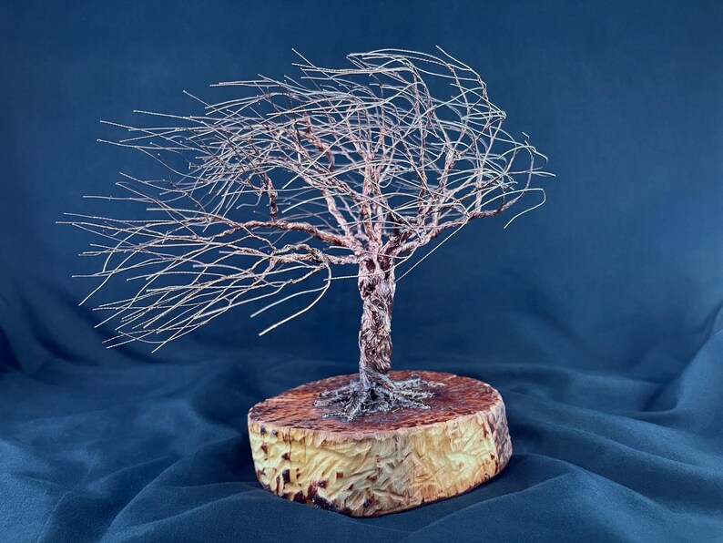 May include: A wire sculpture of a tree with thin, metallic branches extending to one side as if blown by the wind. The trunk is formed from twisted copper-colored wire, and the base is a circular, light-colored wooden slice with a textured edge.