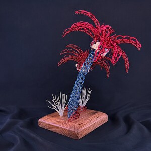 May include: A sculpture of a palm tree crafted from red and blue wire, featuring a textured trunk and fronds. The tree is mounted on a square wooden base, accompanied by two small white wire structures resembling coral or sea plants at the base.