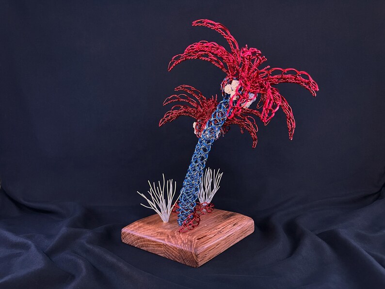 May include: A sculpture of a palm tree crafted from red and blue wire, featuring a textured trunk and fronds. The tree is mounted on a square wooden base, accompanied by two small white wire structures resembling coral or sea plants at the base.