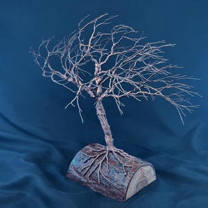 Puede incluir: Una escultura de &aacute;rbol hecha de alambre trenzado de color cobre, con ramas intrincadas que se extienden hacia afuera. El &aacute;rbol est&aacute; anclado a una base hecha de un segmento de tronco de madera natural con corteza y veta visibles.