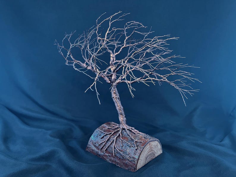 Puede incluir: Una escultura de &aacute;rbol hecha de alambre trenzado de color cobre, con ramas intrincadas que se extienden hacia afuera. El &aacute;rbol est&aacute; anclado a una base hecha de un segmento de tronco de madera natural con corteza y veta visibles.