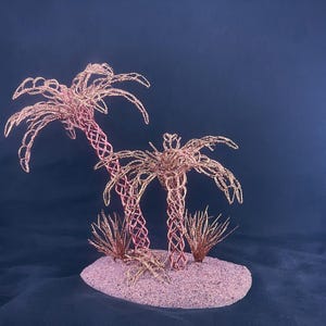 May include: A decorative sculpture featuring two palm trees crafted from thin, copper-colored wire with intricate lattice trunks and fronds. The trees are positioned on a textured, oval-shaped base covered in fine, light pink sand with small wire tufts.