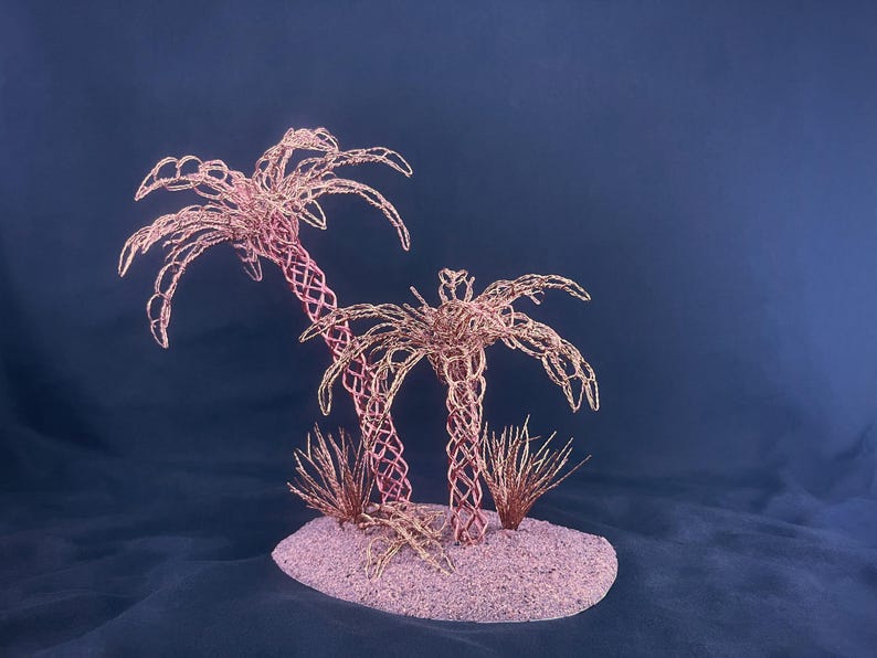 May include: A decorative sculpture featuring two palm trees crafted from thin, copper-colored wire with intricate lattice trunks and fronds. The trees are positioned on a textured, oval-shaped base covered in fine, light pink sand with small wire tufts.