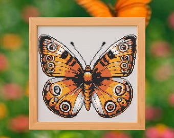 May include: A framed cross-stitch depicting a butterfly with orange, black, and white patterns. The butterfly is set against a white background, within a light brown frame. The background is blurred, featuring green and pink hues.