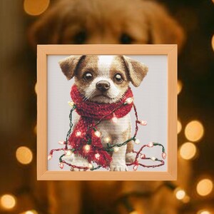 May include: A framed print of a small dog with brown and white fur, wearing a red scarf and wrapped in Christmas lights. The dog is the focal point, set against a neutral background, with a blurred background of another dog.
