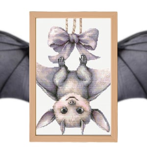 Puede incluir: Un punto de cruz enmarcado de un bebé murciélago colgado boca abajo, con un gran lazo lavanda. El murciélago tiene un cuerpo gris claro, mejillas rosadas y orejas grandes. La obra de arte está en un marco de madera clara.
