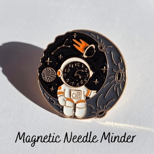 May include: A round magnetic needle minder featuring a space theme with a white and orange astronaut whose helmet faceplate displays a clock face with numbers. The background shows a black starry sky, a grey crescent moon, and a small planet.