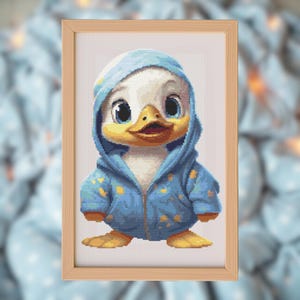 May include: A framed cross-stitch of a cartoon duckling wearing a blue hooded jacket with yellow star patterns. The duckling has a white body, yellow beak and feet, and large blue eyes. The frame is made of light brown wood.