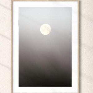 Könnte beinhalten: Ein gerahmter Wandkunstdruck mit einem Vollmond, der in einem vertikalen Farbverlaufshimmel zentriert ist, der von einem hellen Grau oben zu einem tiefen, dunklen Anthrazitschwarz unten übergeht. Der Rahmen besteht aus hellem Holz mit sauberem Finish.