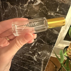 May include: A clear rectangular glass roller bottle with a gold-colored cap. The word grace is etched in a cursive script font on the front surface of the glass bottle. The bottle has a smooth, transparent finish and a metallic gold lid.