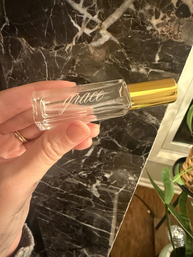 May include: A clear rectangular glass roller bottle with a gold-colored cap. The word grace is etched in a cursive script font on the front surface of the glass bottle. The bottle has a smooth, transparent finish and a metallic gold lid.