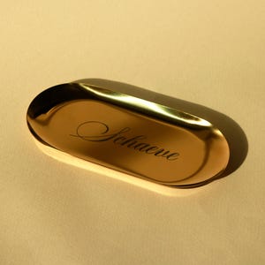 Minimalist oval metal trinket tray in gold finish on white background, modern jewelry dish for rings, bracelets, necklaces or keys.