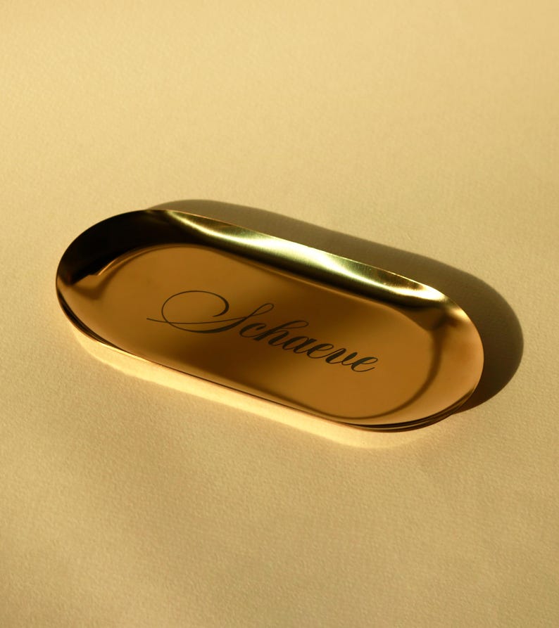 Minimalist oval metal trinket tray in gold finish on white background, modern jewelry dish for rings, bracelets, necklaces or keys.