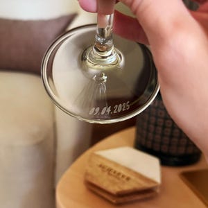 Op de afbeelding: Een helder wijnglas met de datum 09.04.2025 gegraveerd op de bodem. Het glas wordt omhoog gehouden, waarbij de steel en de kelk te zien zijn. Een klein decoratief element is zichtbaar in de kelk. De achtergrond omvat een houten onderzetter en een decoratieve fles.