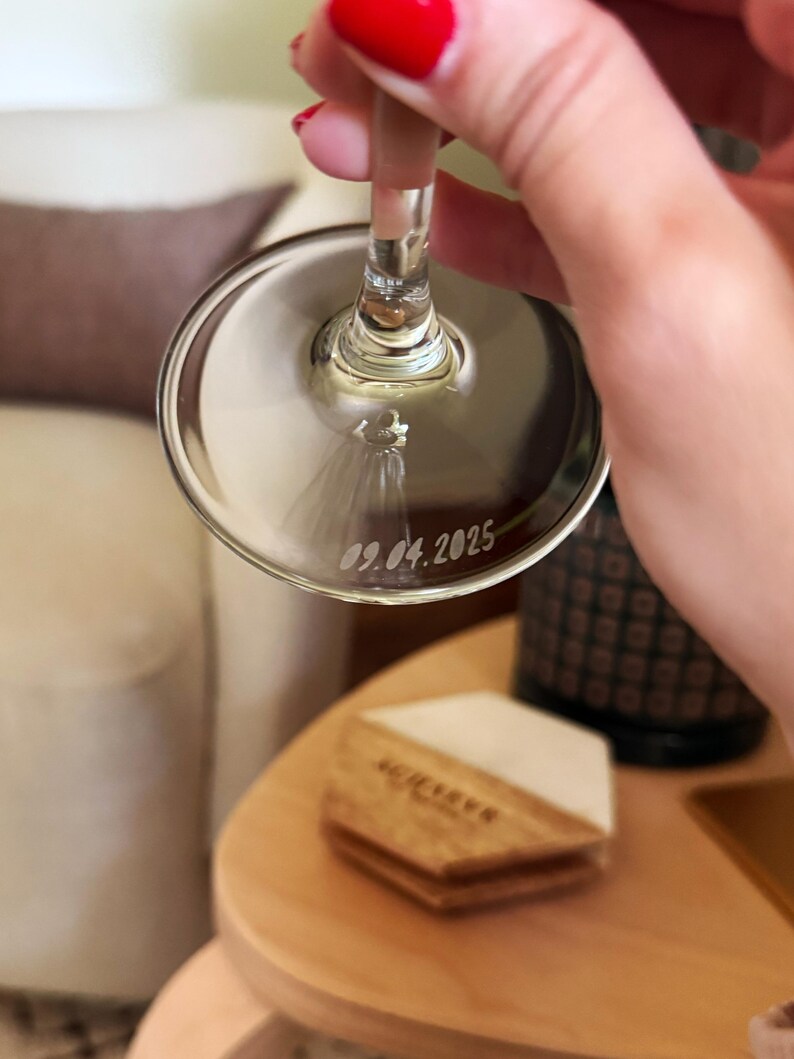 Op de afbeelding: Een helder wijnglas met de datum 09.04.2025 gegraveerd op de bodem. Het glas wordt omhoog gehouden, waarbij de steel en de kelk te zien zijn. Een klein decoratief element is zichtbaar in de kelk. De achtergrond omvat een houten onderzetter en een decoratieve fles.