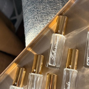 May include: A collection of rectangular clear glass perfume bottles with gold-colored caps. Each bottle features a white script name printed on the front, including Katherine, Erin, and Linda, with a small white heart icon on each gold cap.