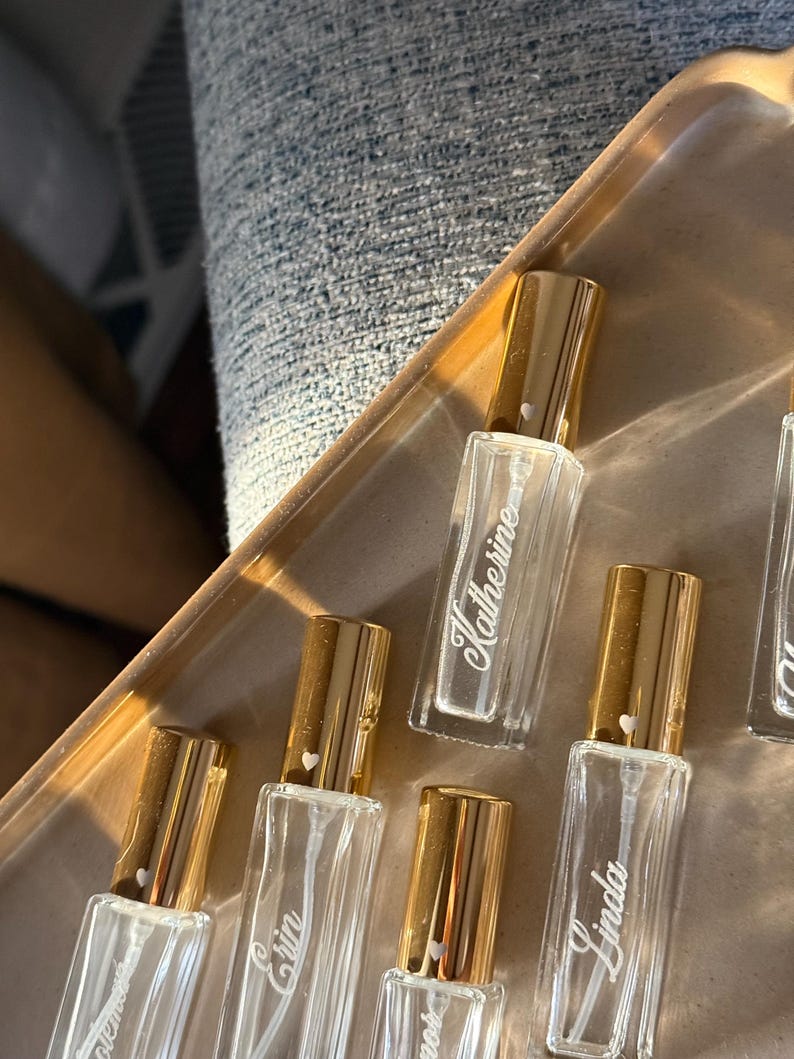 May include: A collection of rectangular clear glass perfume bottles with gold-colored caps. Each bottle features a white script name printed on the front, including Katherine, Erin, and Linda, with a small white heart icon on each gold cap.