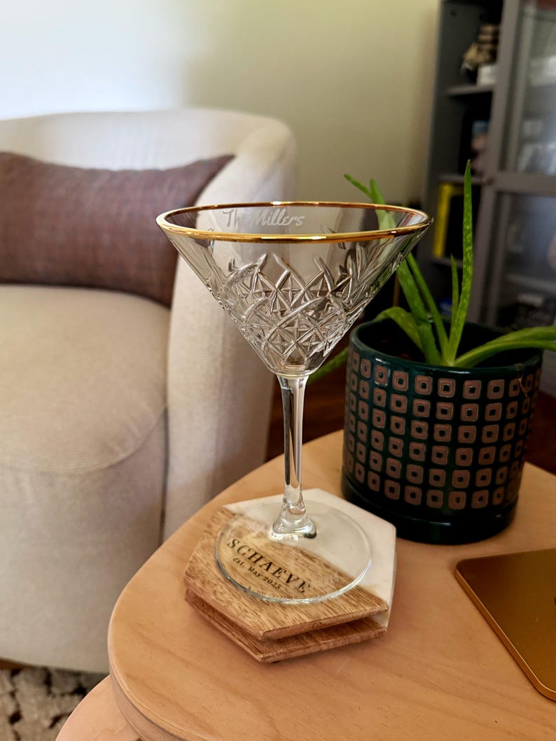 Op de afbeelding: Een helder kristallen martini glas met een gouden rand en de gegraveerde tekst "The Millers". Het glas staat op een houten en marmeren onderzetter met de tekst "SCHAEVE" en "Est. May 2015".