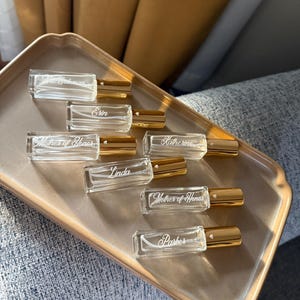 May include: A collection of seven rectangular glass perfume roller bottles with gold caps. Each bottle features white engraved text including names like Erin, Katherine, Linda, Parker, and the title Mother of Honor, with a small heart icon near the cap.