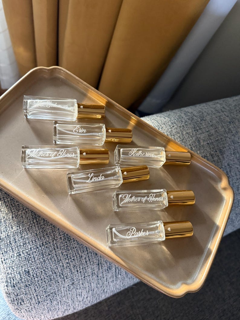 May include: A collection of seven rectangular glass perfume roller bottles with gold caps. Each bottle features white engraved text including names like Erin, Katherine, Linda, Parker, and the title Mother of Honor, with a small heart icon near the cap.
