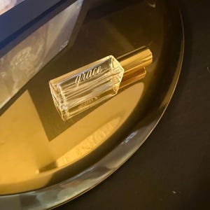 May include: A rectangular clear glass perfume bottle with a gold-toned cylindrical cap. The word grace is etched in a cursive script font on the front face of the glass bottle. The bottle is empty and features a thick, transparent base.