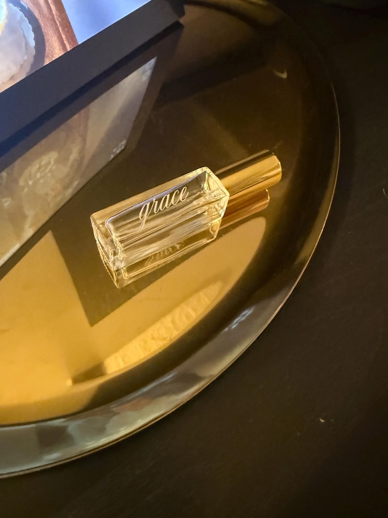 May include: A rectangular clear glass perfume bottle with a gold-toned cylindrical cap. The word grace is etched in a cursive script font on the front face of the glass bottle. The bottle is empty and features a thick, transparent base.