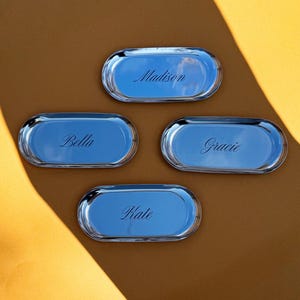 Op de afbeelding: Vier ovale zilverkleurige metalen dienbladen met een gepolijste, reflecterende afwerking. Elk dienblad is voorzien van een unieke naam gegraveerd in een cursief lettertype: Madison, Bella, Gracie en Kate. De dienbladen liggen in een groepje.