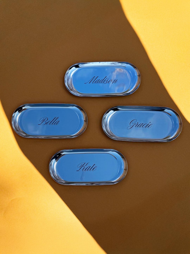 Op de afbeelding: Vier ovale zilverkleurige metalen dienbladen met een gepolijste, reflecterende afwerking. Elk dienblad is voorzien van een unieke naam gegraveerd in een cursief lettertype: Madison, Bella, Gracie en Kate. De dienbladen liggen in een groepje.