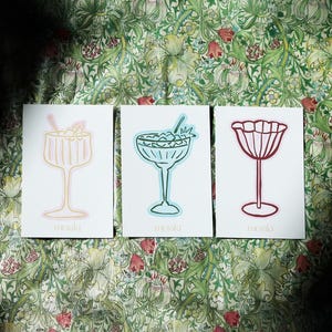 Op de afbeelding: Drie witte kaarten met cocktailillustraties in roze, blauwe en rode contouren. Elke kaart heeft een ander drankontwerp, met het woord "meraki" onderaan gedrukt. De kaarten zijn geplaatst tegen een bloemig patroon.