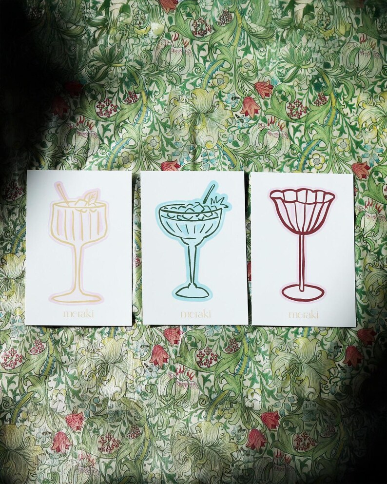 Op de afbeelding: Drie witte kaarten met cocktailillustraties in roze, blauwe en rode contouren. Elke kaart heeft een ander drankontwerp, met het woord "meraki" onderaan gedrukt. De kaarten zijn geplaatst tegen een bloemig patroon.