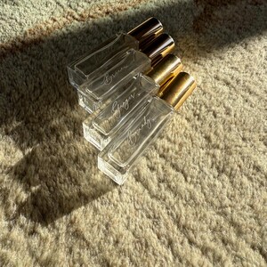 May include: Four rectangular clear glass perfume bottles with gold-colored caps arranged in a diagonal stack. Each bottle features white cursive script text on the front, displaying the names Bruna, Ginger, and Brandy, with one bottle appearing blank.