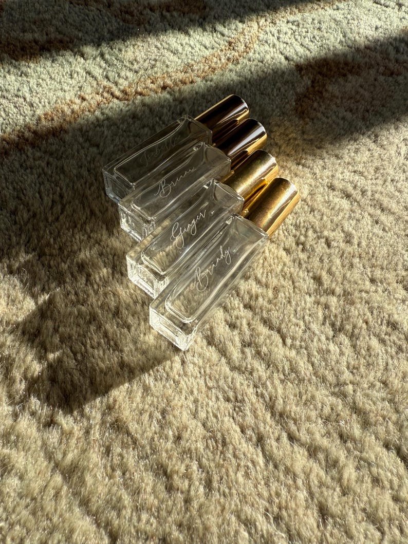 May include: Four rectangular clear glass perfume bottles with gold-colored caps arranged in a diagonal stack. Each bottle features white cursive script text on the front, displaying the names Bruna, Ginger, and Brandy, with one bottle appearing blank.