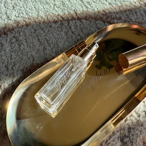 May include: A rectangular clear glass perfume bottle with a gold spray nozzle and a matching gold cap, resting on an oval gold-toned metal tray. The bottle features the word Ginger printed in white script, while the tray displays the word Stephens.