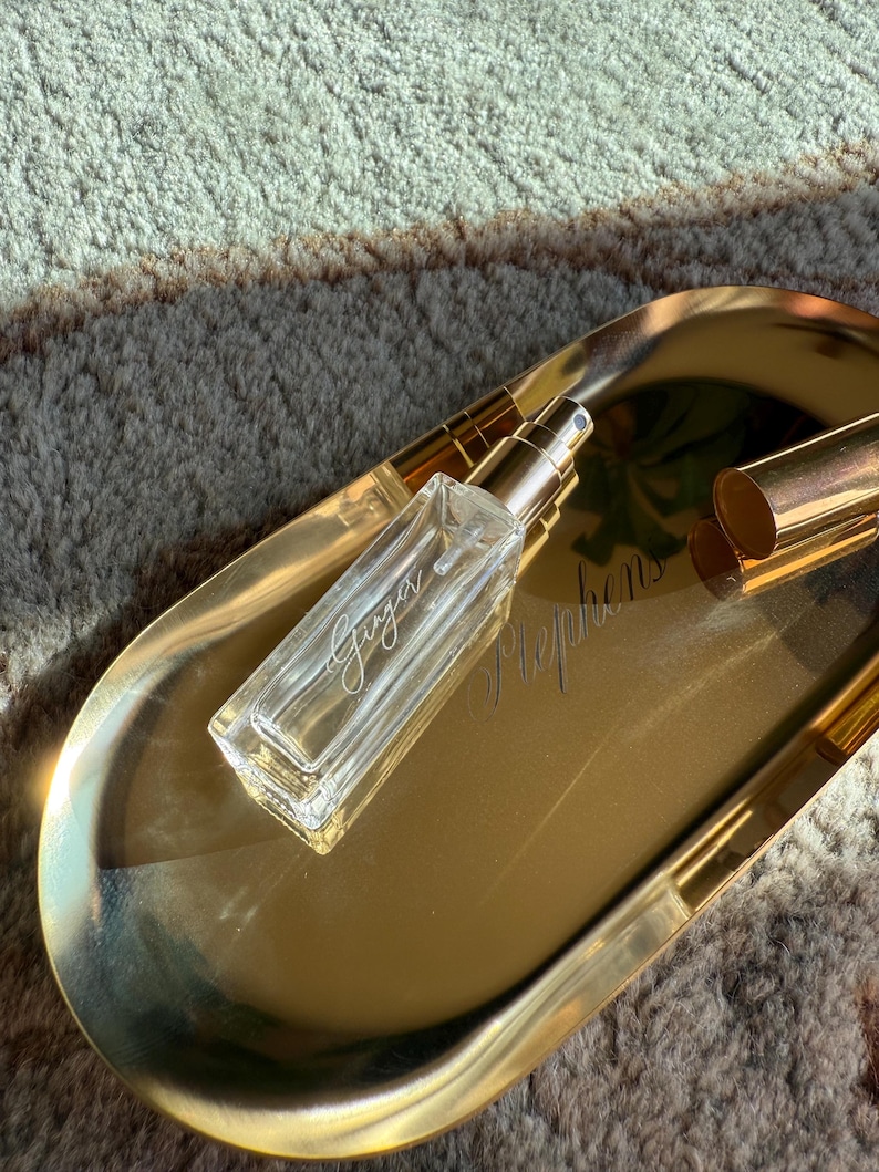 May include: A rectangular clear glass perfume bottle with a gold spray nozzle and a matching gold cap, resting on an oval gold-toned metal tray. The bottle features the word Ginger printed in white script, while the tray displays the word Stephens.