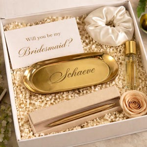Op de afbeelding: Een geschenkdoos met een kaart met de tekst Will you be my Bridesmaid?, een gouden ovaal dienblad gegraveerd met de naam Schaeve, een witte satijnen scrunchie, een glazen flesje met gouden dop, een gouden pen in een etui en een perzikkleurige roos.