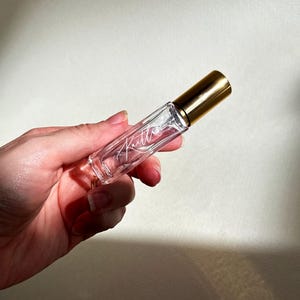 May include: A clear glass roller bottle with a gold-colored cap. The bottle features the name Kaitlin written in a white cursive script on the front surface. The container is cylindrical and transparent, showing the empty interior space clearly.
