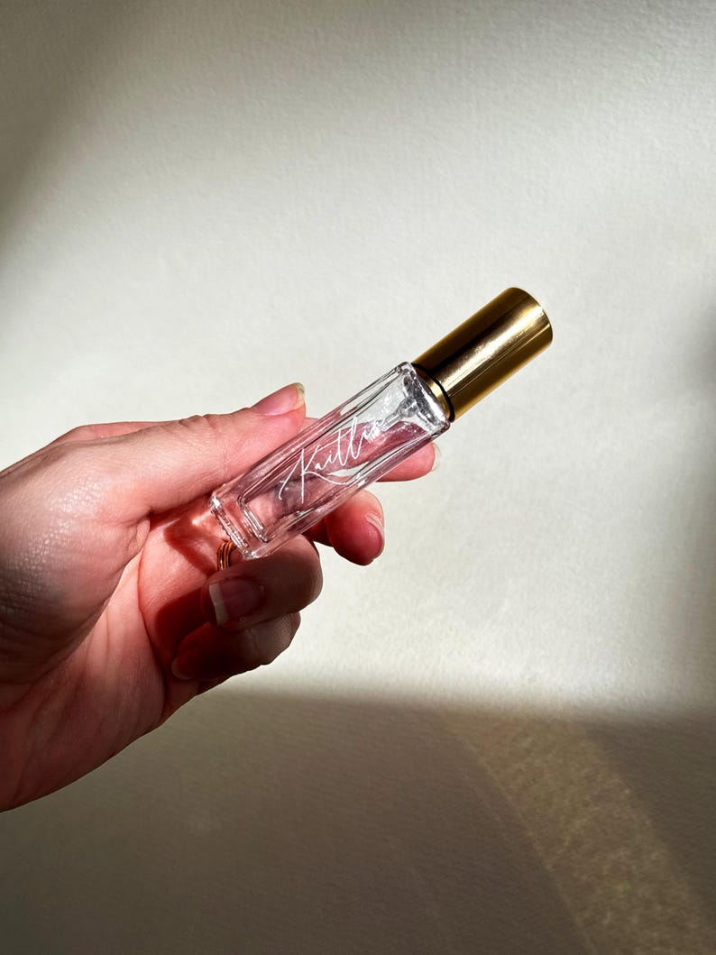 May include: A clear glass roller bottle with a gold-colored cap. The bottle features the name Kaitlin written in a white cursive script on the front surface. The container is cylindrical and transparent, showing the empty interior space clearly.