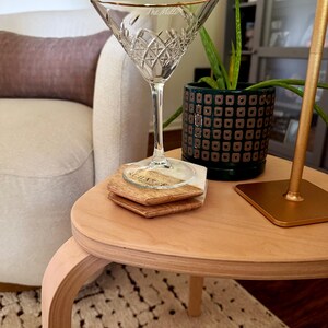 Op de afbeelding: Een helder kristallen martiniglas met een gouden rand, op een houten tafel. Het glas heeft een ruitpatroon en de tekst "The Miller". Twee houten onderzetters met "Sugar" zijn eronder gestapeld. Een plant is zichtbaar.